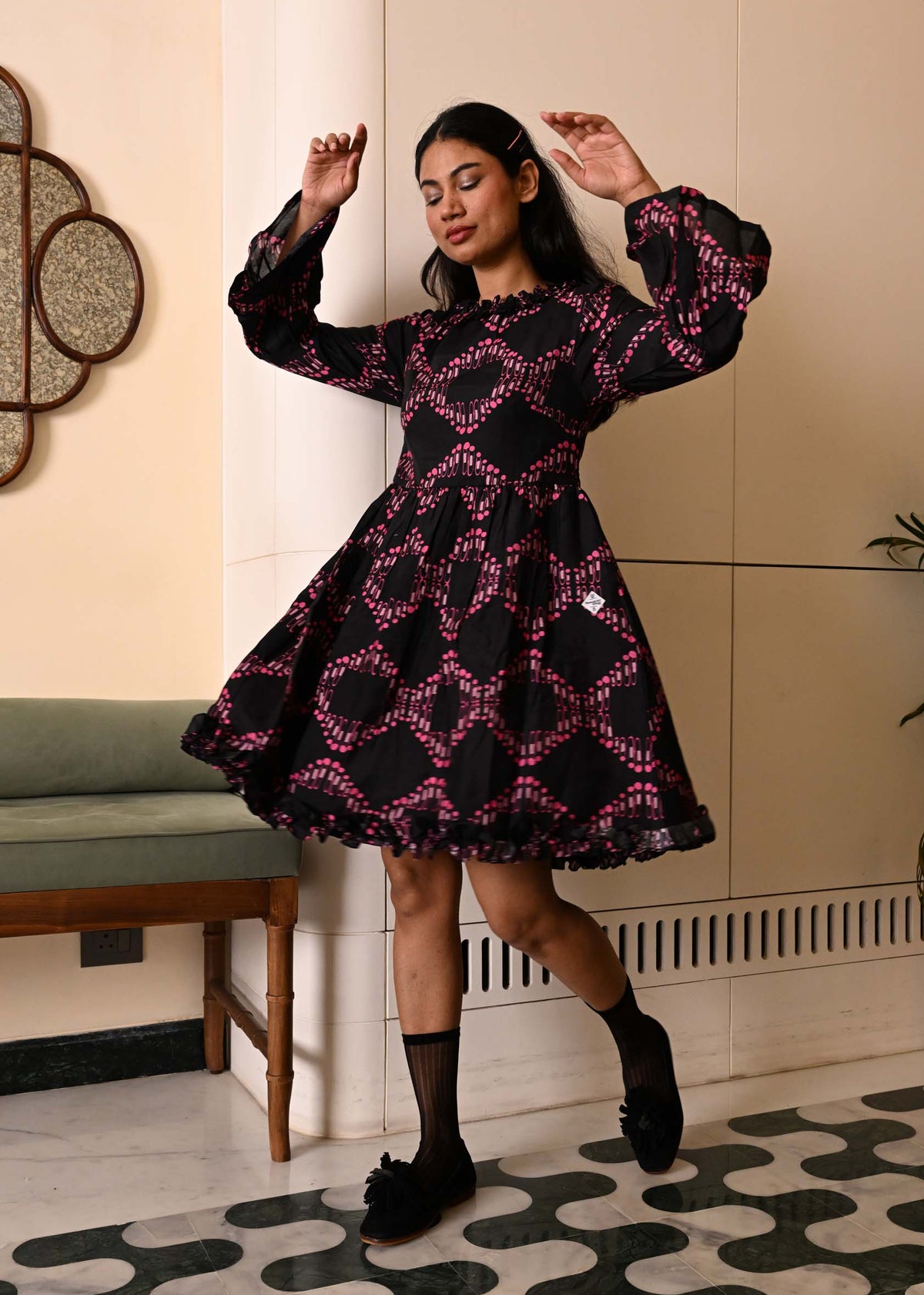A woman modeling a black long-sleeve mini dress with a pink geometric pattern, standing in a modern indoor setting with a patterned floor and a green bench in the background.