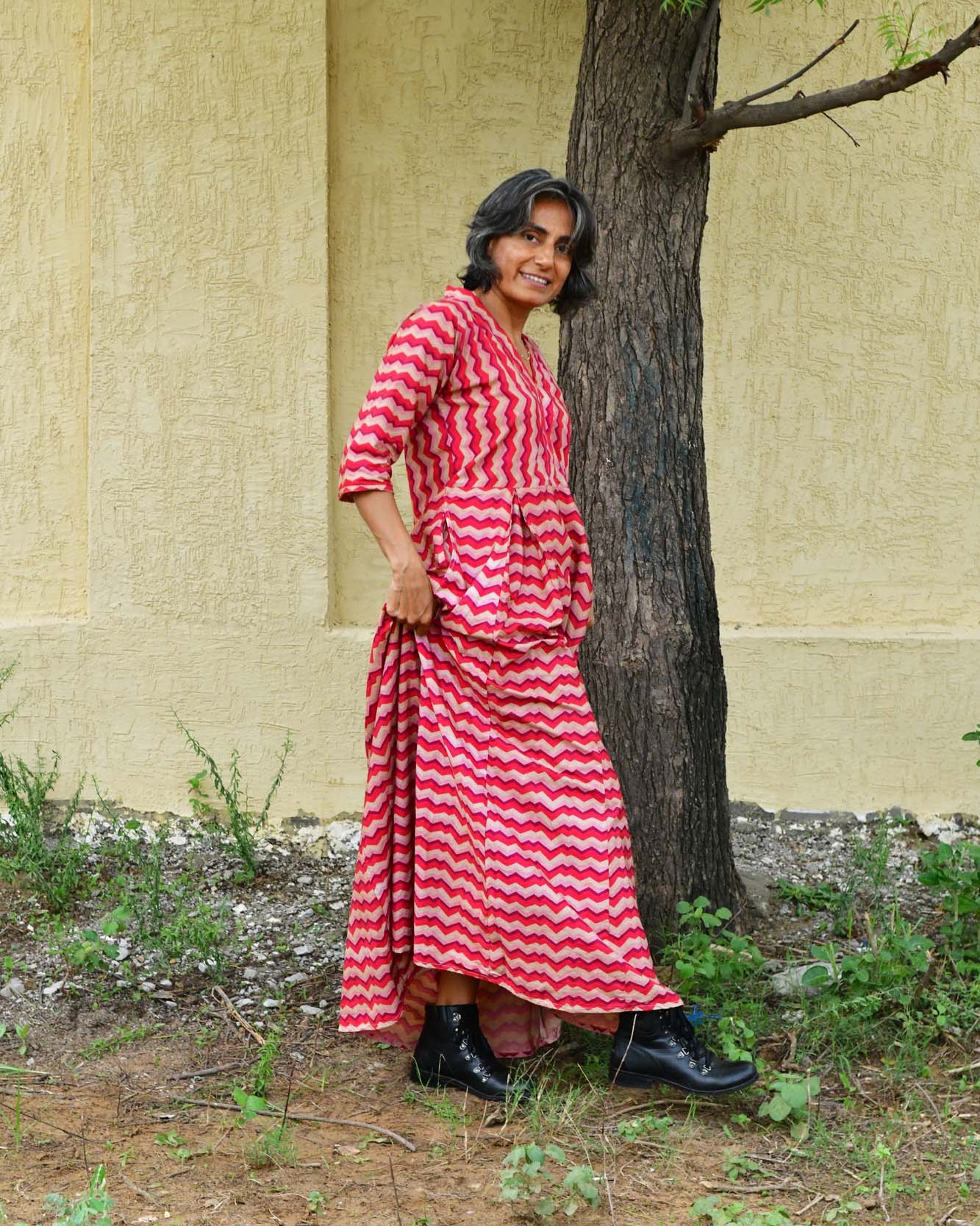 Woman in a red and white striped dress standing next to a tree against a beige wall.
