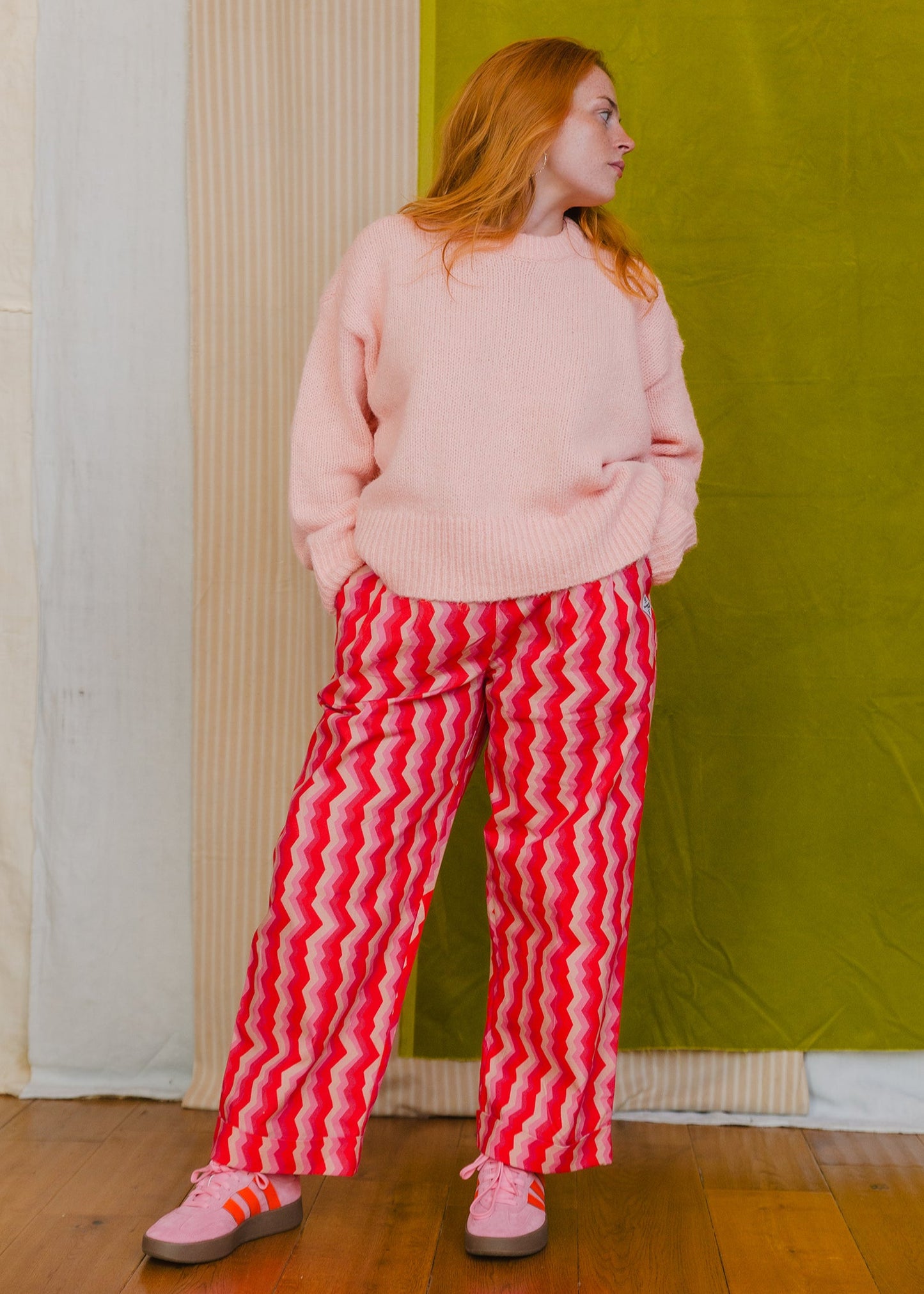 Person wearing a pink sweater and red striped pants standing against a green wall.