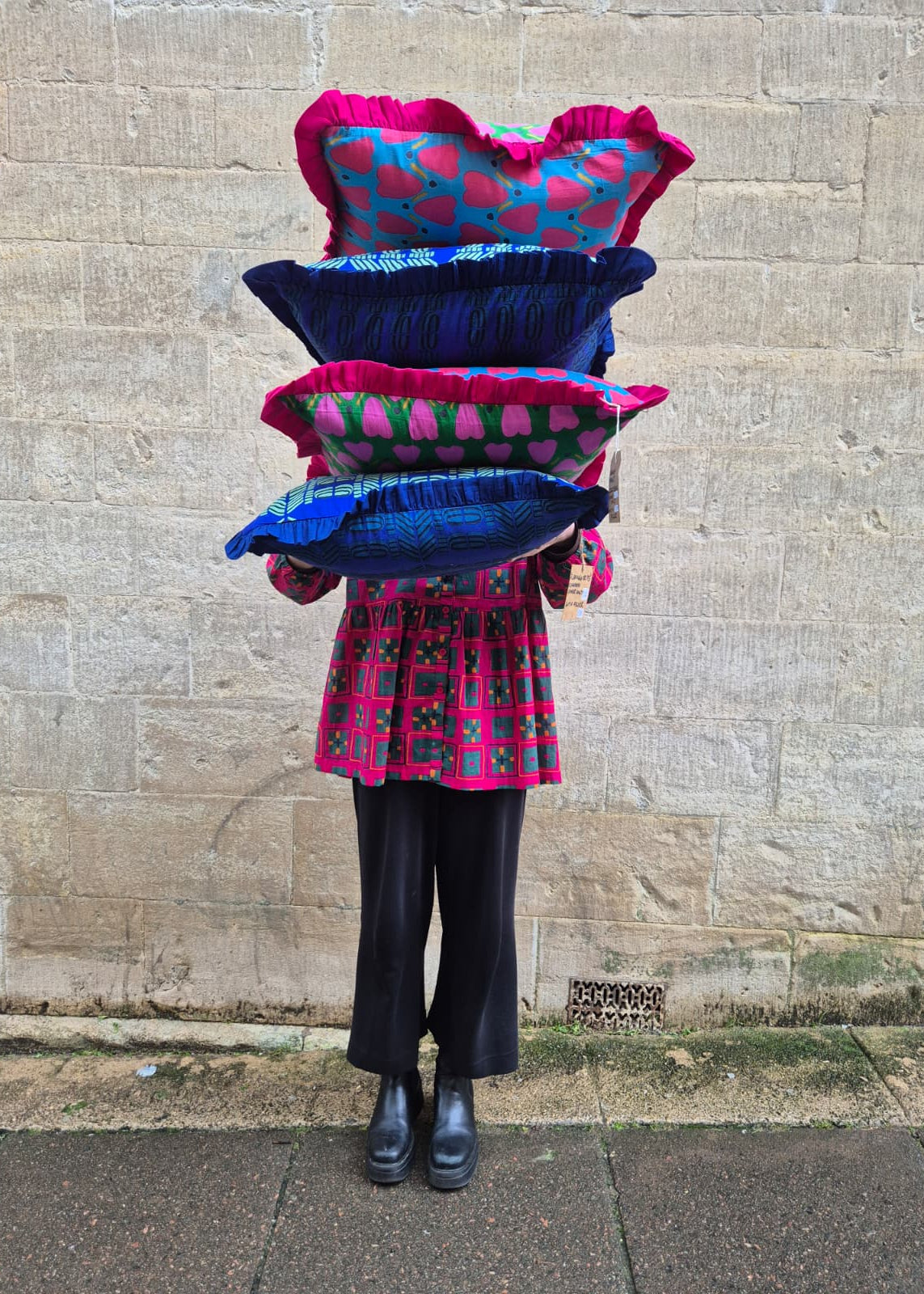 Person holding a stack of colorful pillows against a stone wall.