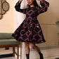A woman modeling a black long-sleeve mini dress with a pink geometric pattern, standing in a modern indoor setting with a patterned floor and a green bench in the background.
