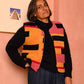 A woman stands in a pink room wearing a colorful geometric patterned knit vest over a black turtleneck, loose dark blue jeans, and black sneakers.