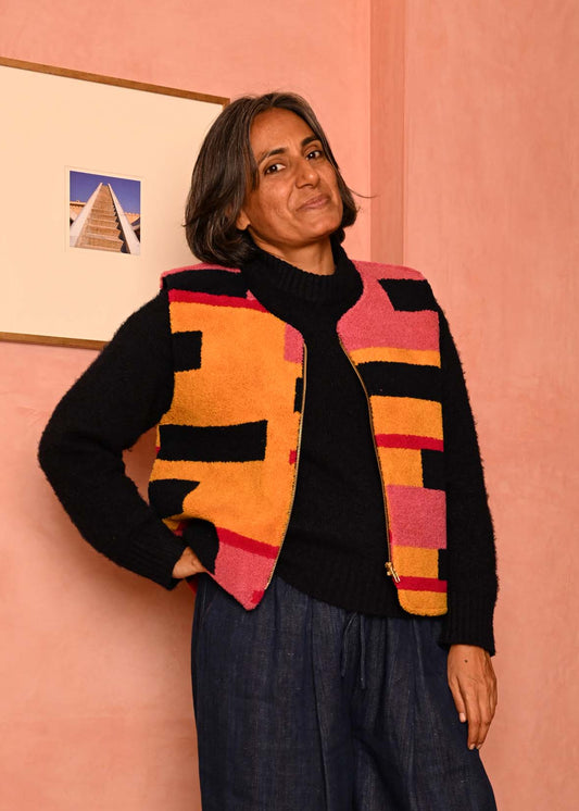 A woman stands in a pink room wearing a colorful geometric patterned knit vest over a black turtleneck, loose dark blue jeans, and black sneakers.