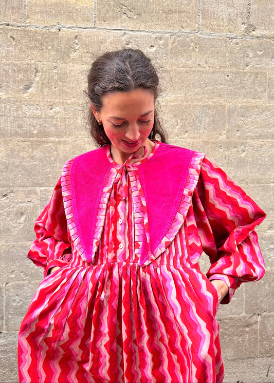 Woman wearing a pink and red patterned dress against a stone wall with long pointy collar.